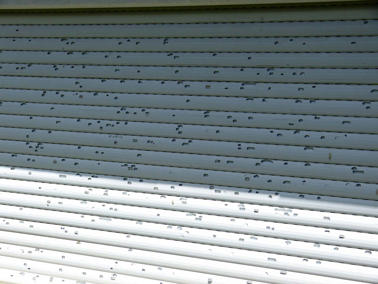 A hail-damaged roller shutter with multiple holes showing the extent of hail damage to the building